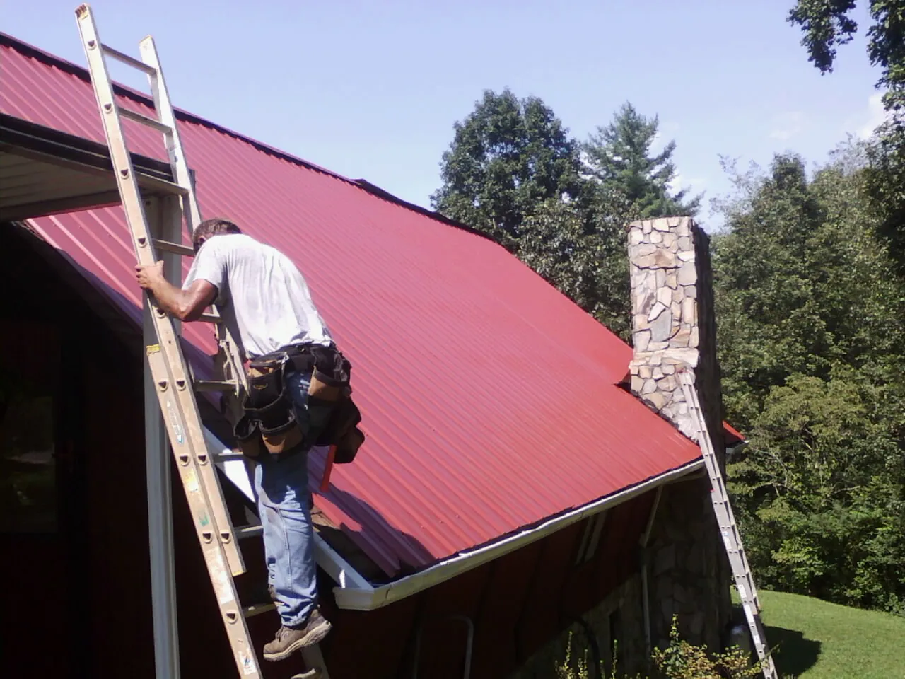 Roofer on a ladder beside a red metal roof and stone chimney — Soffit & Fascia Repair by Roofing Brothers in Chester Gap